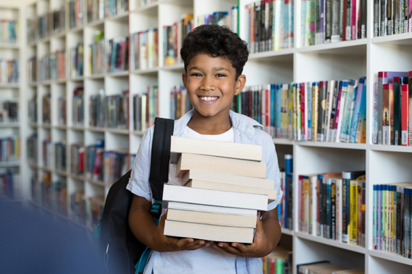 Boy poses with backpack and a stack of books in front of bookshelf