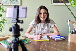 A teenager wearing glasses writes in a notebook and looks into a cell phone on a tripod