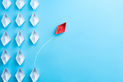 A red paper boat veering away from a set of white paper boats on a blue background
