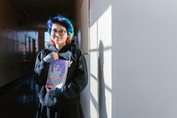 A teenager with glasses and colorful hair standing in a hallway holding notebooks